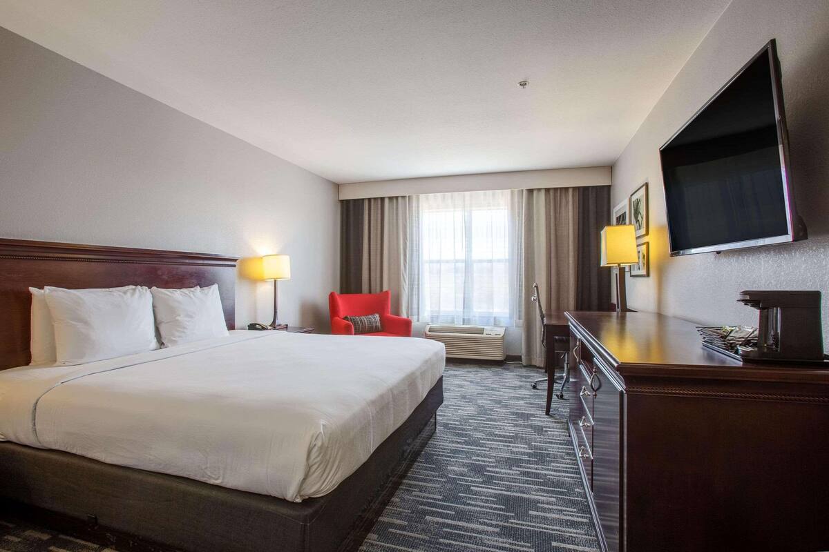 A hotel room with a large bed, a desk with a flat-screen TV, a red chair, and a window with curtains.