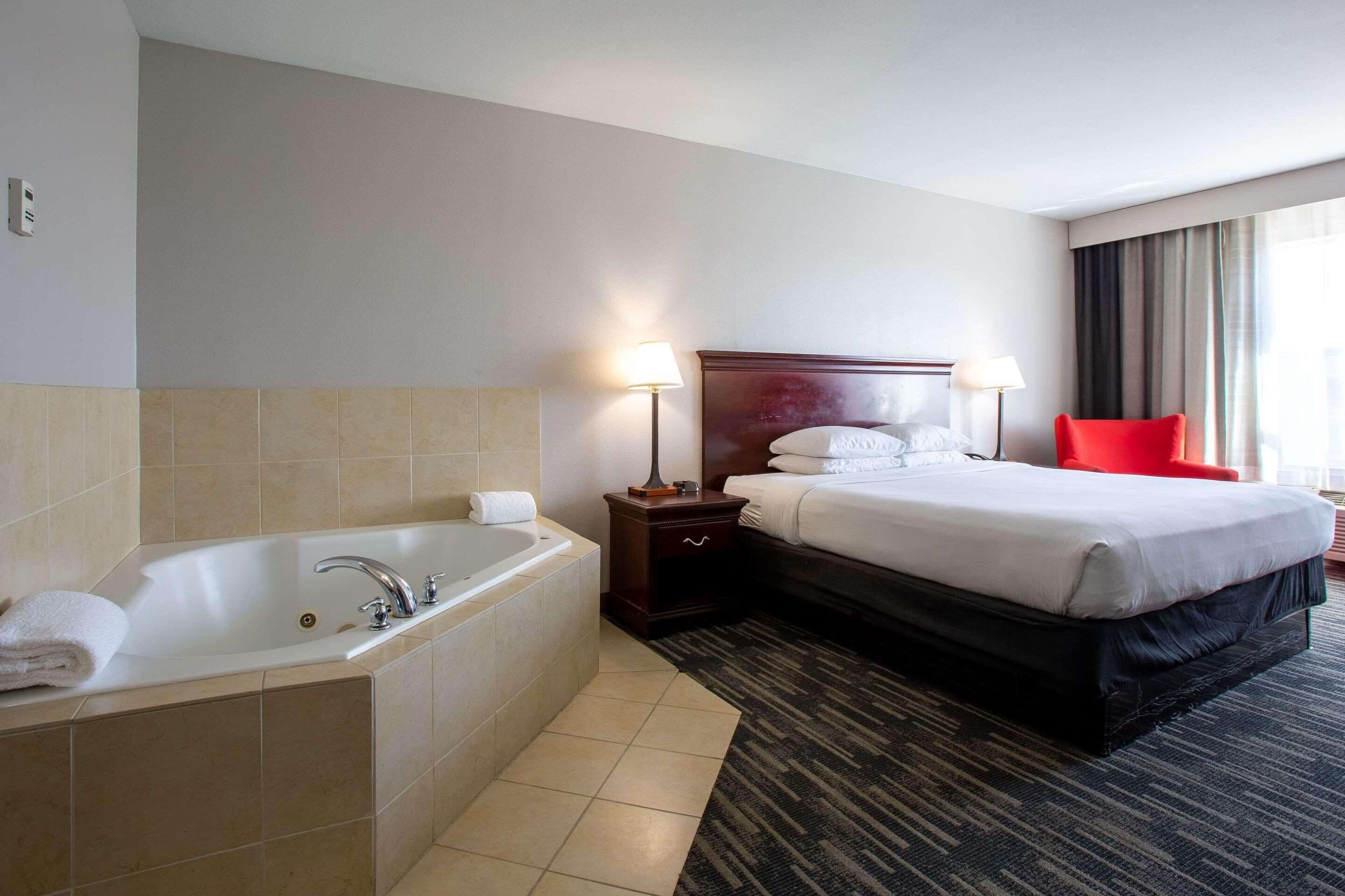 Ein Hotelzimmer mit einer großen Badewanne, einem ordentlich bezogenen Bett, einem roten Sessel und einem Nachttisch mit Lampen.
