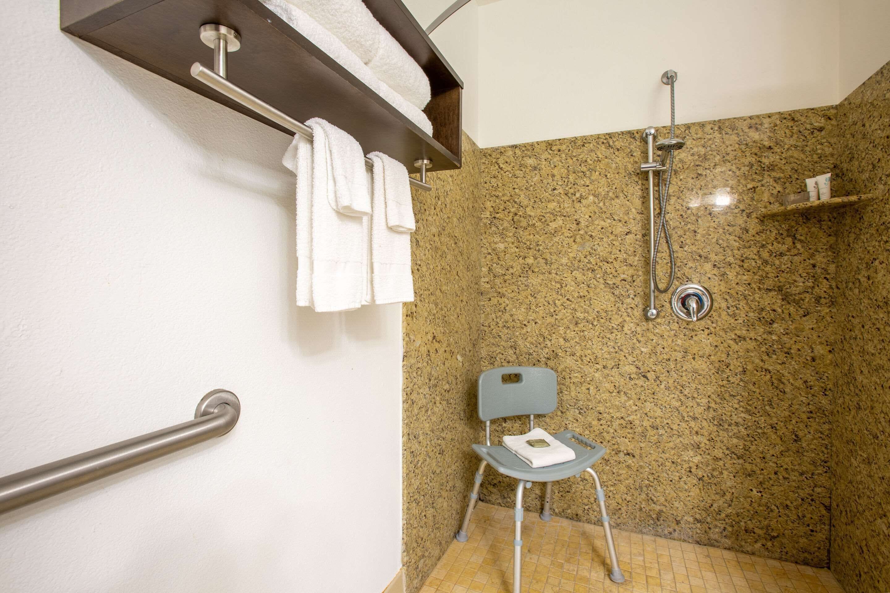 A bathroom with a shower, a towel rack, a chair, and a handrail.