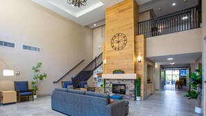 A spacious lobby with a high ceiling, a staircase, a large blue sofa, a wooden wall clock, and a fireplace. - Country Inn & Suites by Radisson, Tucson City Center, AZ (Tucson)