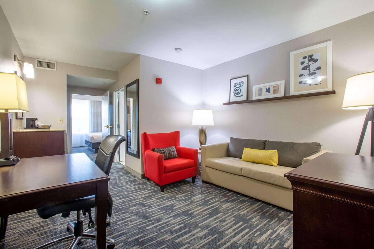A hotel room with a wooden desk, a red armchair, a beige sofa, and a patterned carpet.