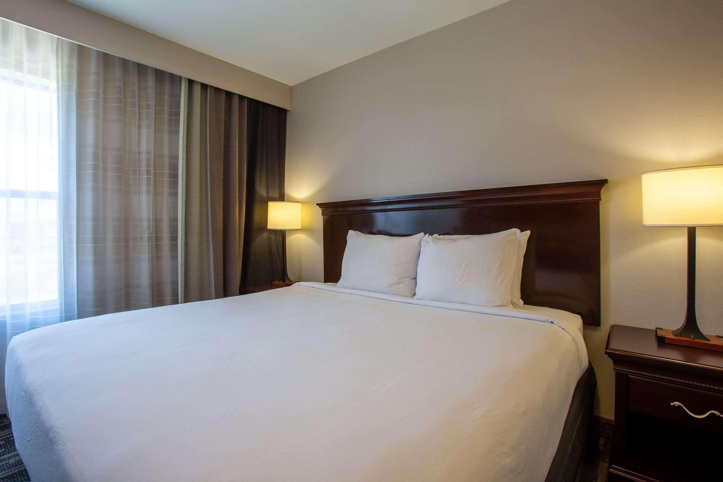 Ein Hotelzimmer mit einem großen Bett, zwei Nachttischlampen, einem Holz-Kopfteil und einem Fenster mit Vorhängen.
