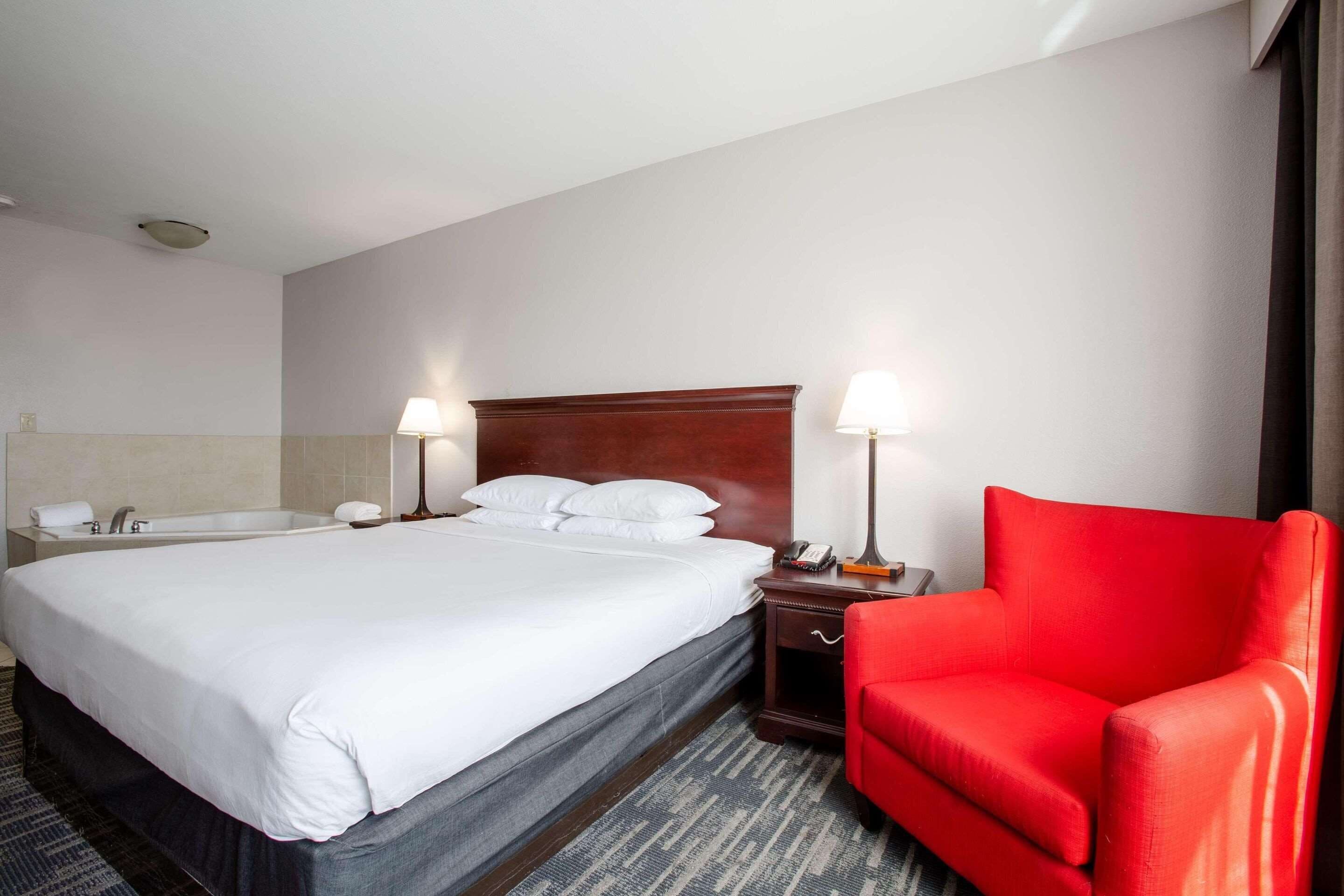 A hotel room with a large bed, a red chair, a nightstand with a lamp, and a bathroom with a bathtub and toilet.