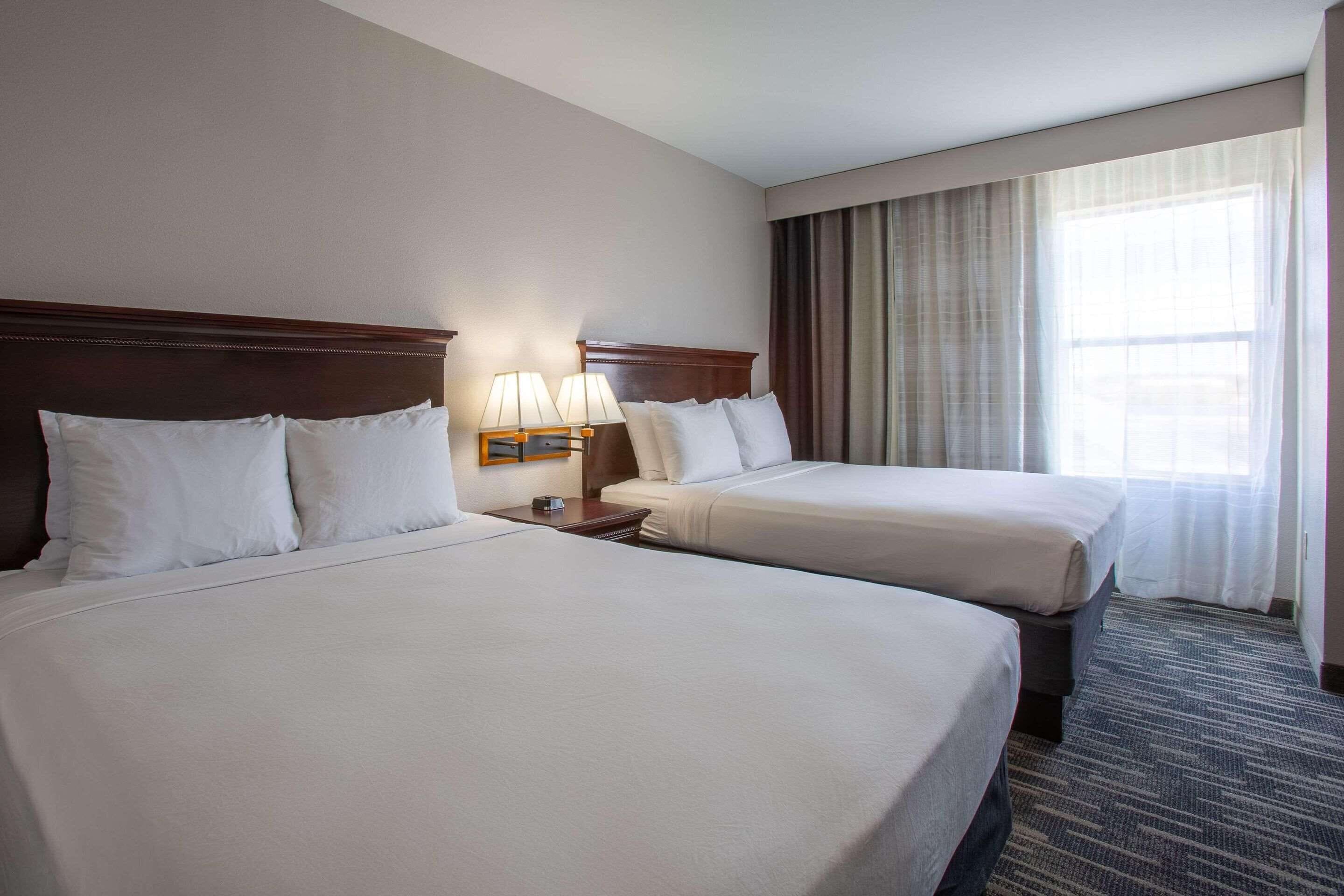 A hotel room with two beds, a nightstand with lamps, a window with curtains, and a carpeted floor.