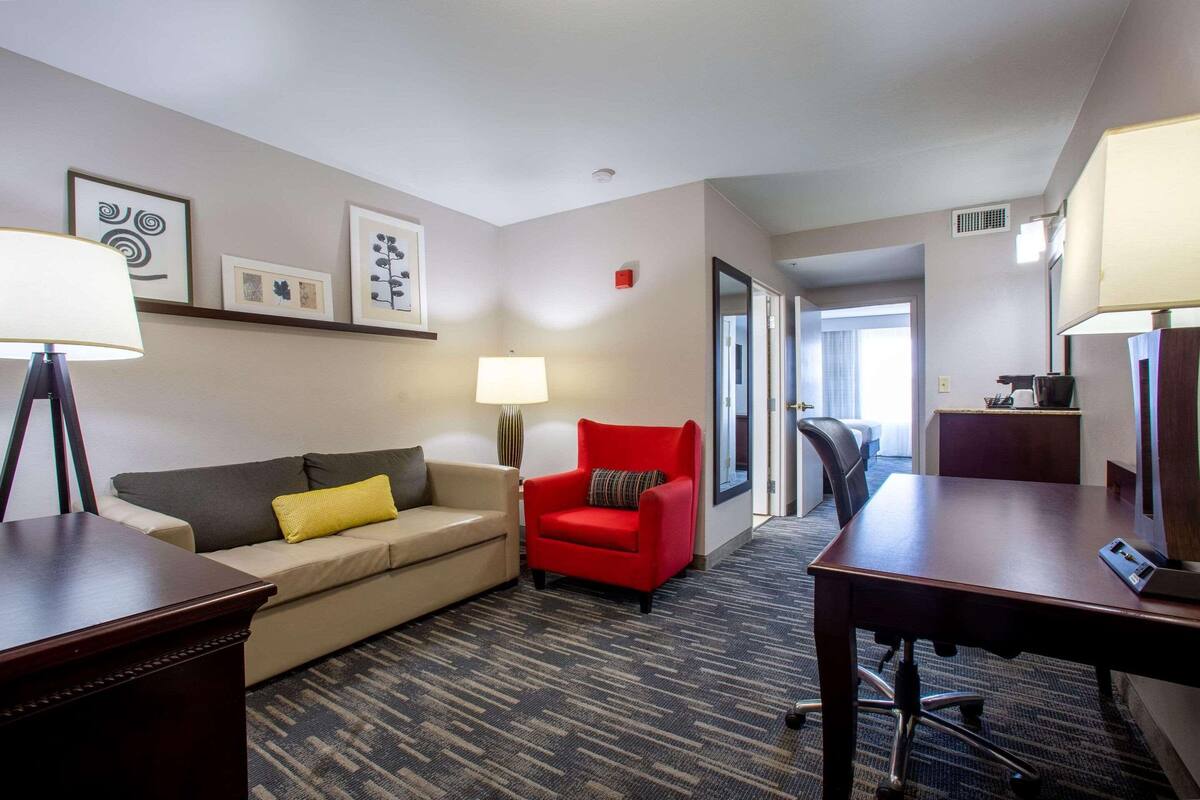 A hotel room with a sofa, a red armchair, a desk, and a lamp. There are framed pictures on the wall and a carpeted floor.