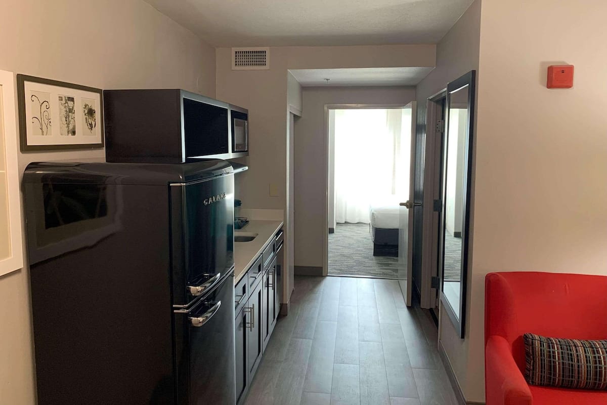 A compact hotel room with a kitchenette, a red armchair, and a bed with a window view.