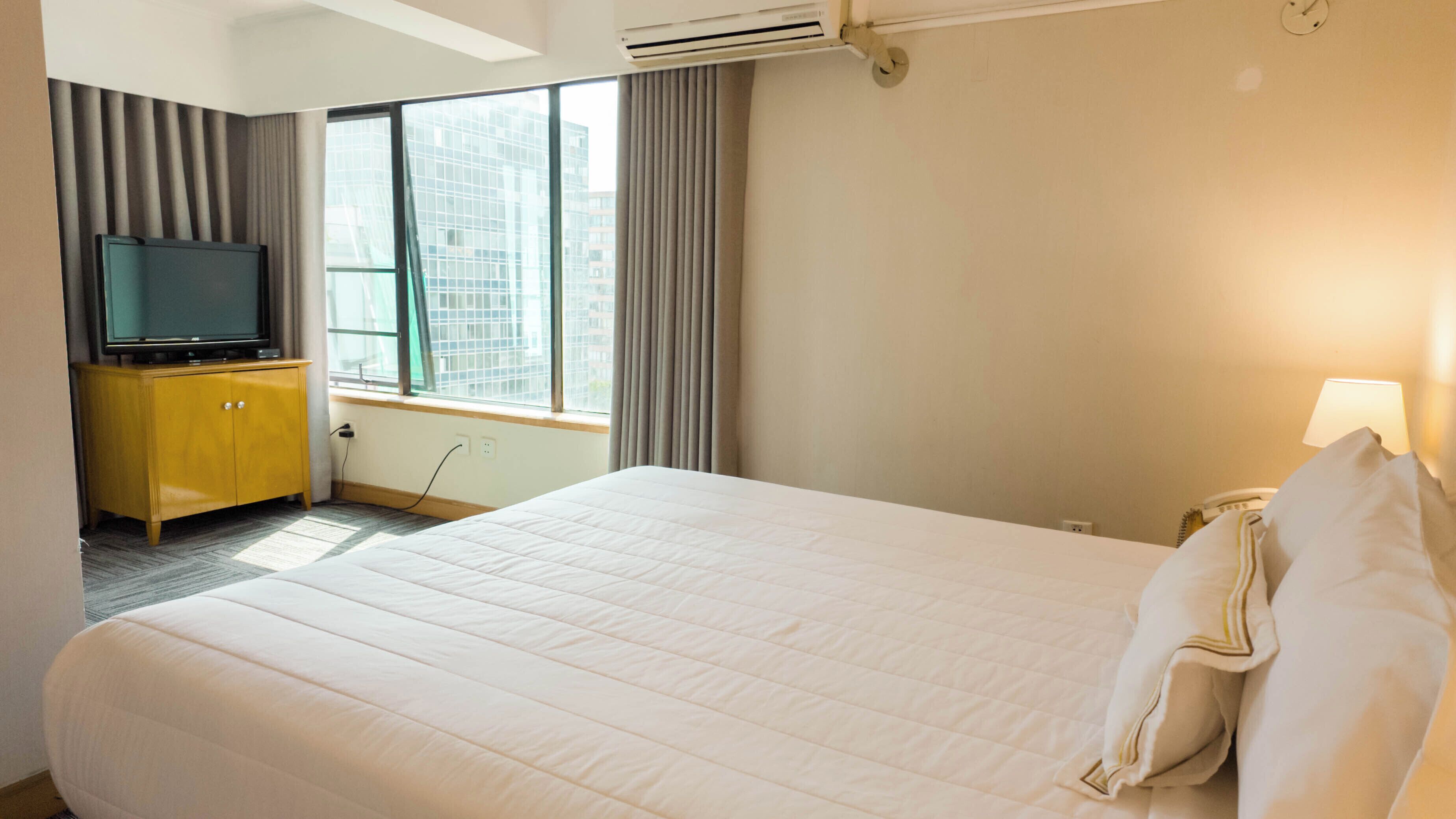 A hotel room with a large bed, a television on a wooden cabinet, a window with curtains, and a bedside lamp.