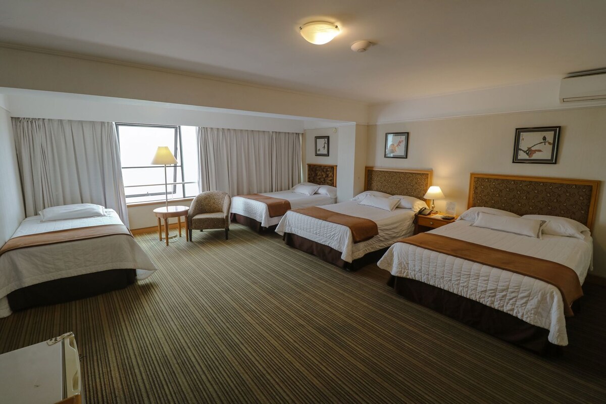 A hotel room with three beds, a chair, a lamp, and a window with curtains.