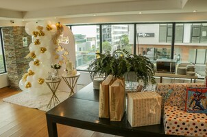 Un interior moderno con una zona de recepción que incluye un arco de globos, una planta en maceta y una mesa con bolsas de regalo y paquetes envueltos.