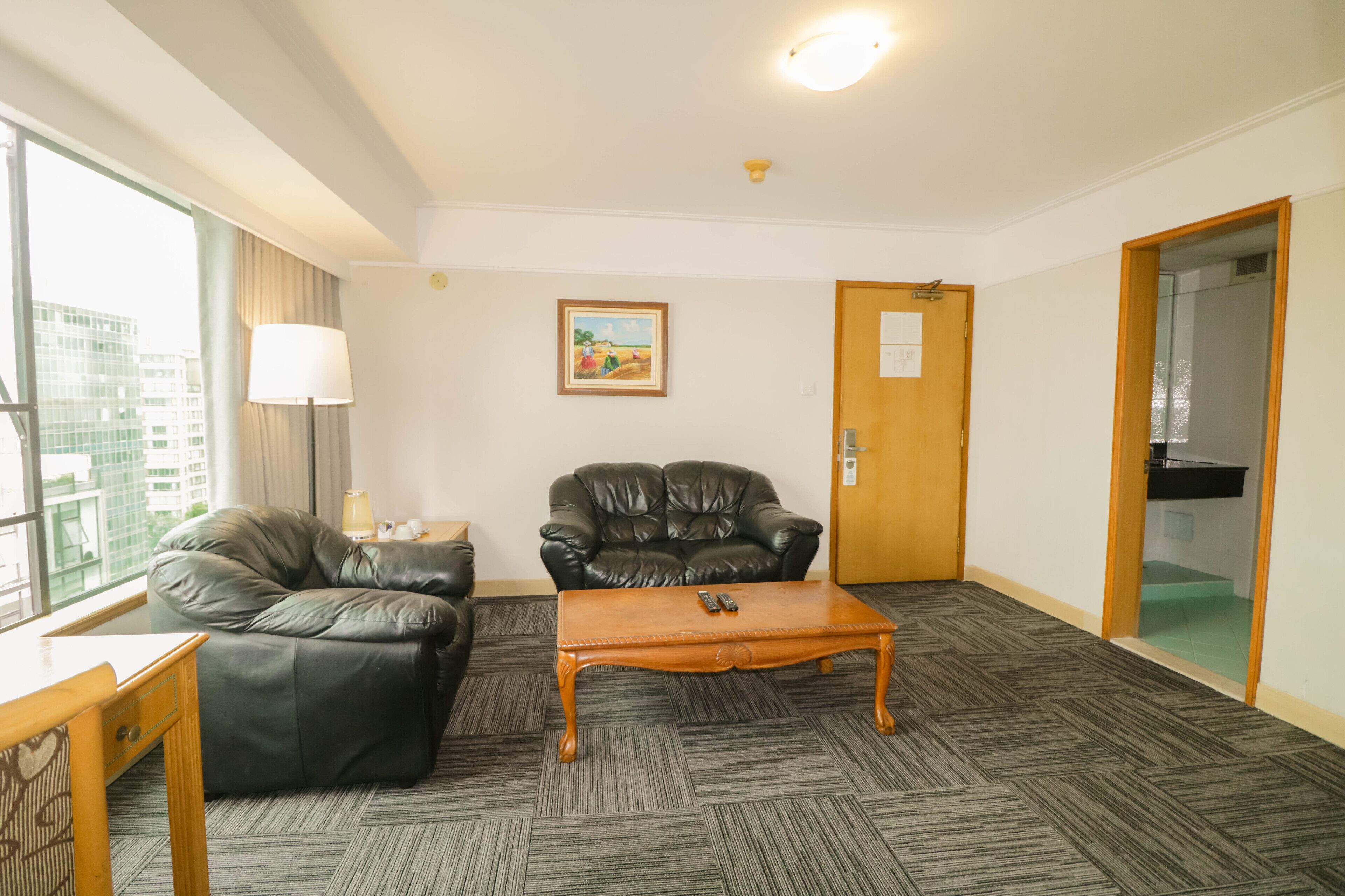 A hotel room with a leather sofa, a wooden coffee table, a desk, a lamp, a painting on the wall, and a door leading to a bathroom.