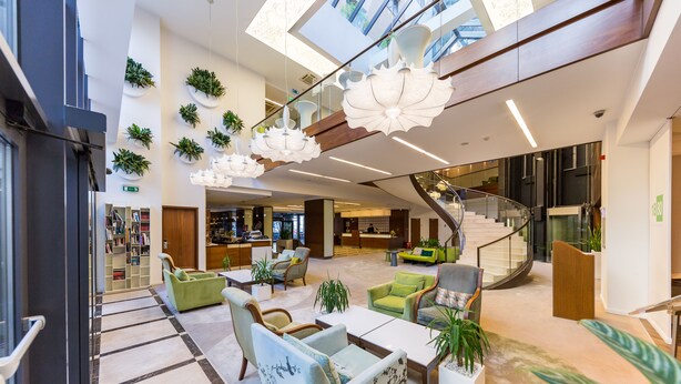 A spacious lobby with a modern design, featuring a central seating area with sofas and armchairs, a staircase, and a variety of potted plants. - Capital Plaza Hotel (Bucharest)