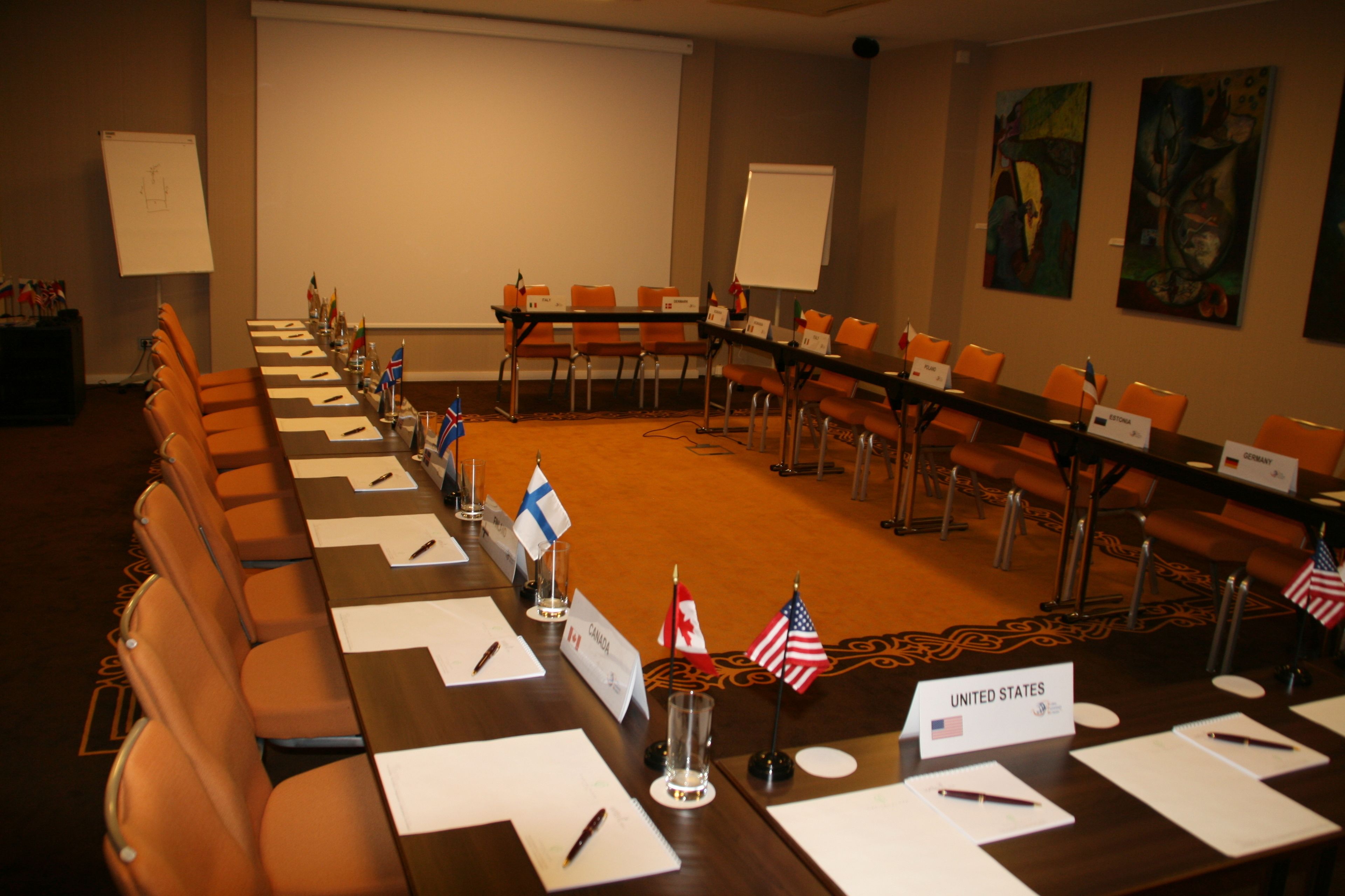 Une salle de conférence avec une longue table, des chaises, des plaques nominatives et des drapeaux.