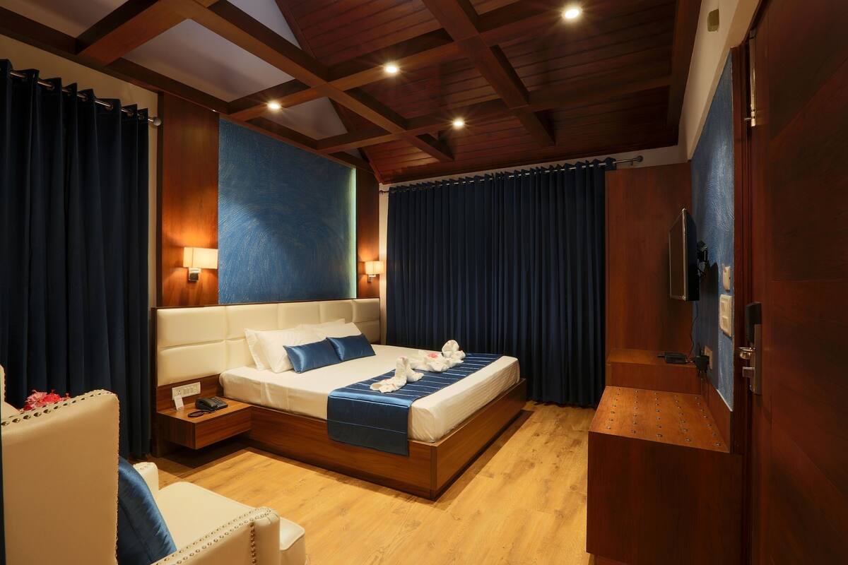 A modern hotel room with a wooden ceiling, a large bed with a blue bedspread, a nightstand, and a seating area with a chair.