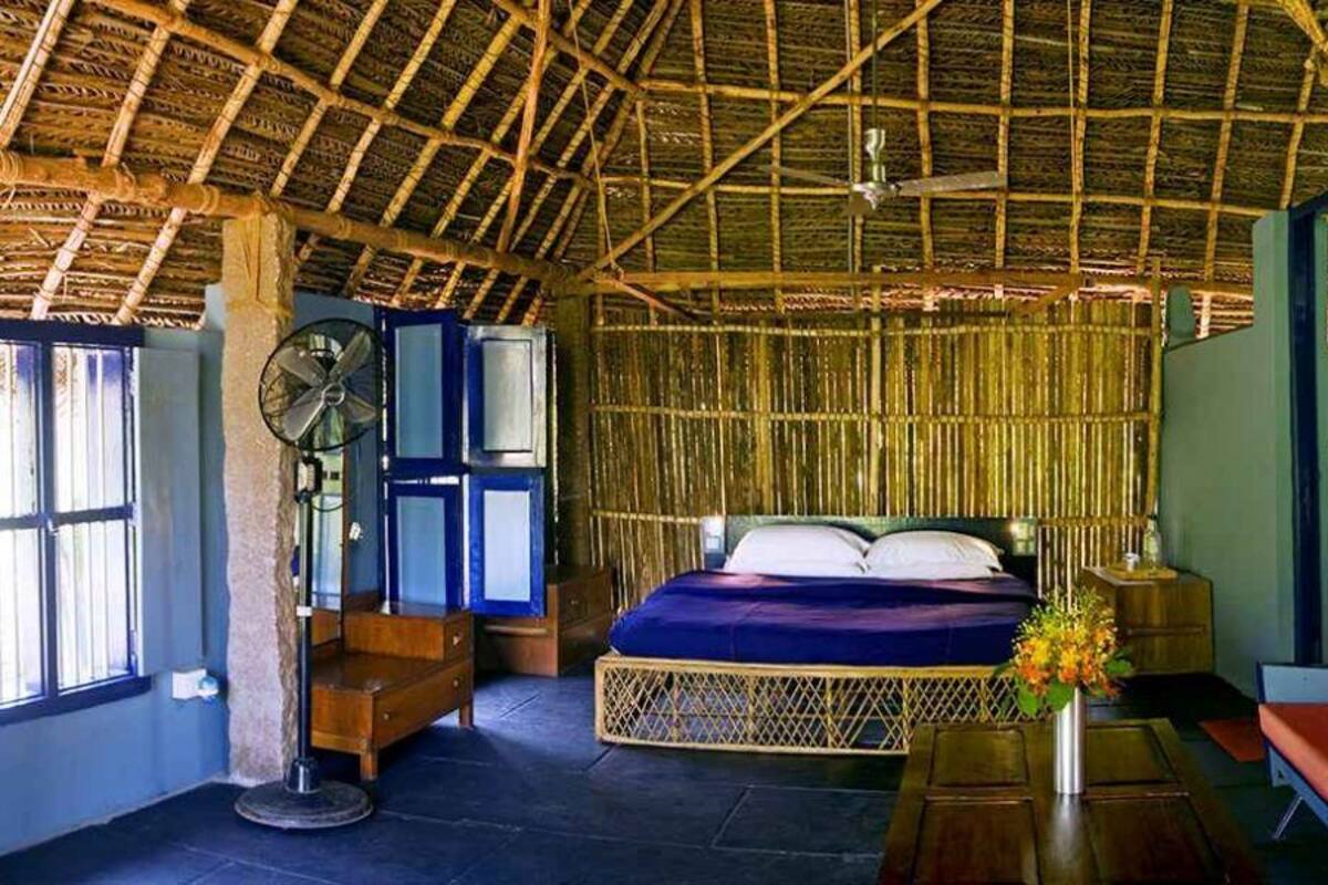 A room with a thatched roof, a bed with a blue mattress, a wooden bench, a fan, and a table with a vase of flowers.