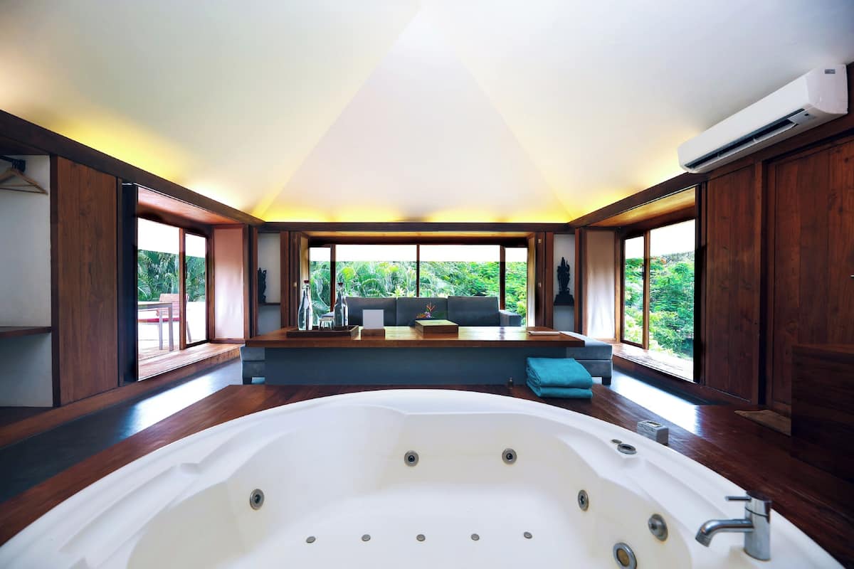 A spacious bathroom with a large bathtub, wooden flooring, and large windows offering a view of greenery.