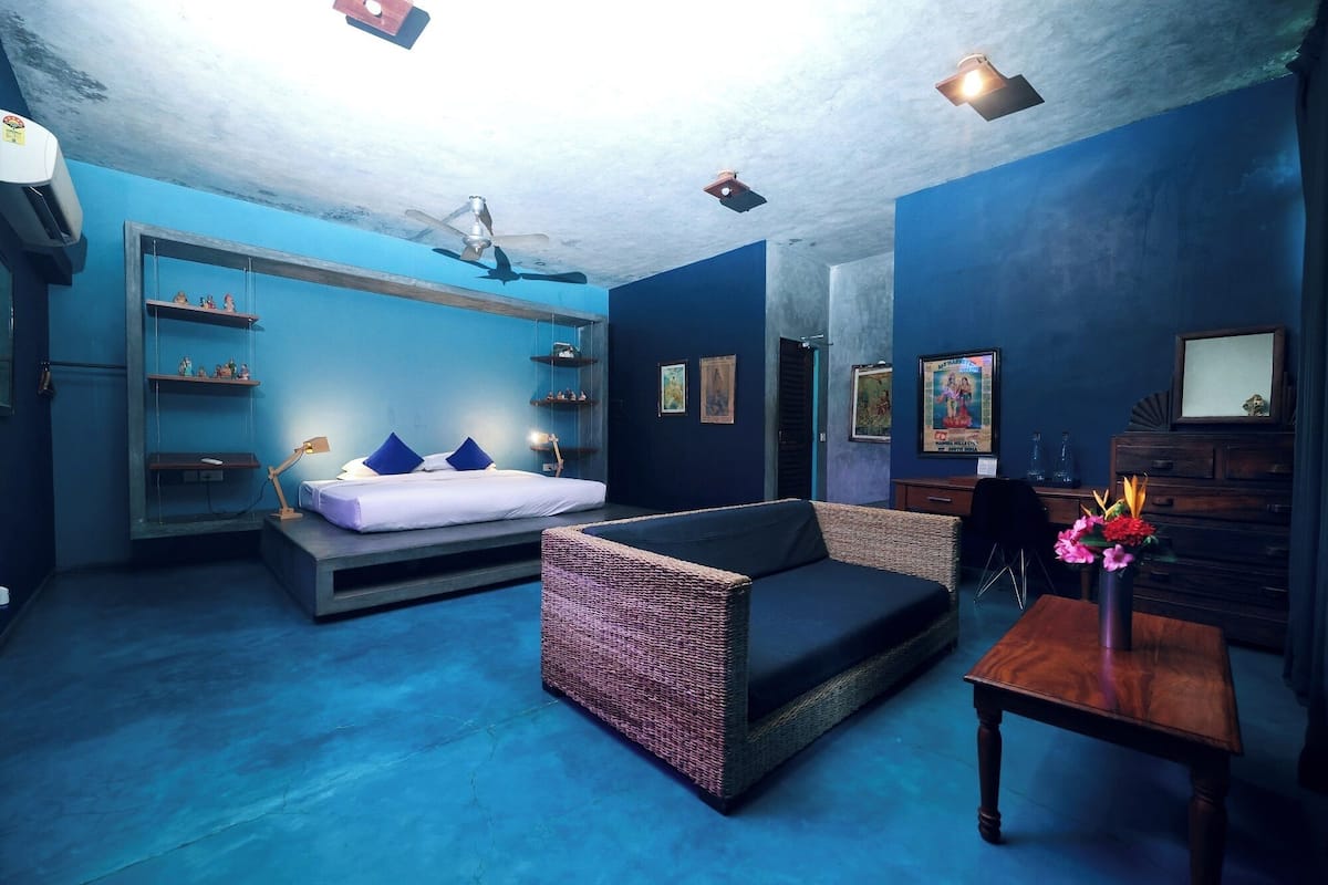 A modern room with a blue color scheme, featuring a bed, a sofa, a small table, and wall-mounted shelves.