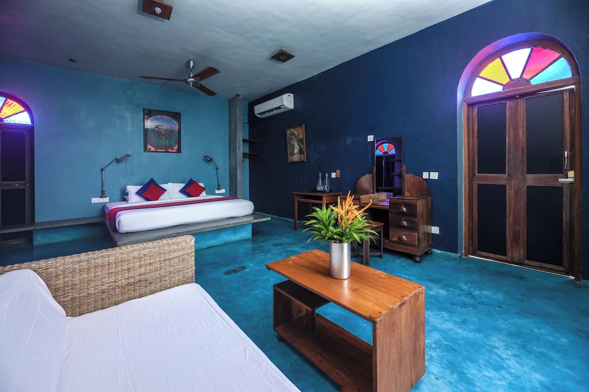 A room with a bed, a wooden table with a plant, a ceiling fan, and a large arched window with stained glass.