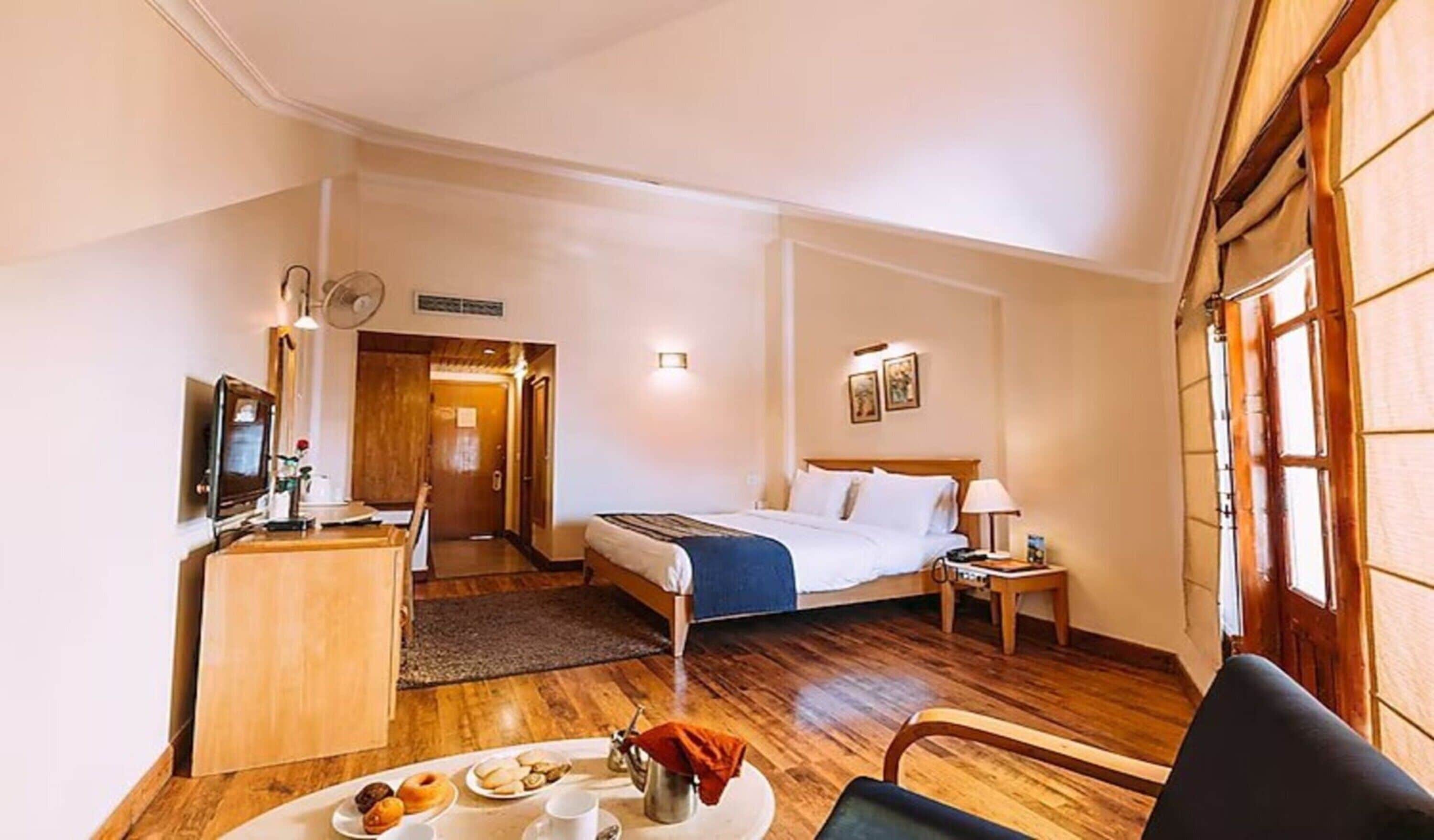 A hotel room with a bed, bedside table, lamp, and a dining area with a table set for breakfast.