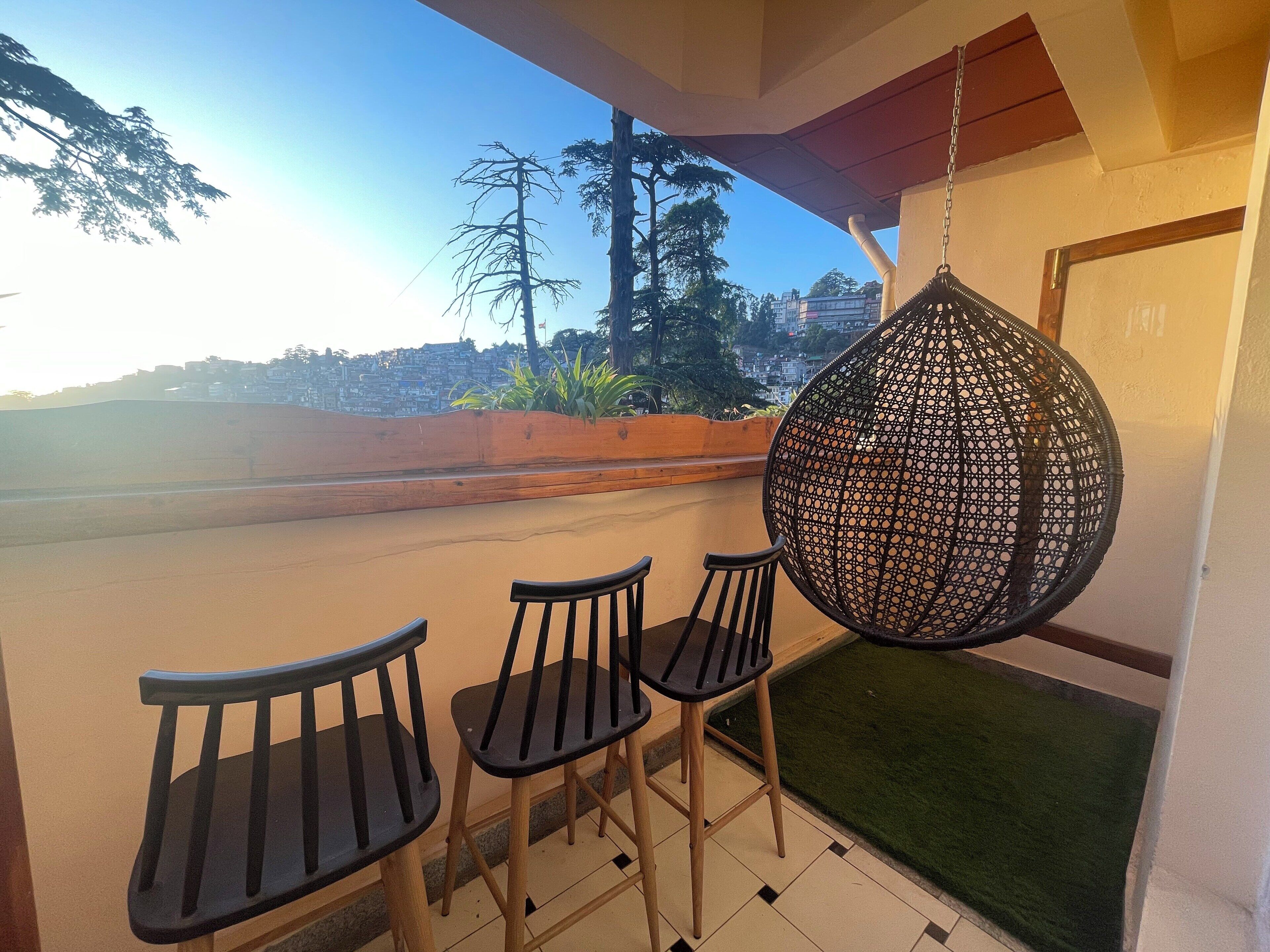 Un balcón con tres sillas de madera y una silla colgante de ratán, con vista a un paisaje urbano de edificios y árboles.