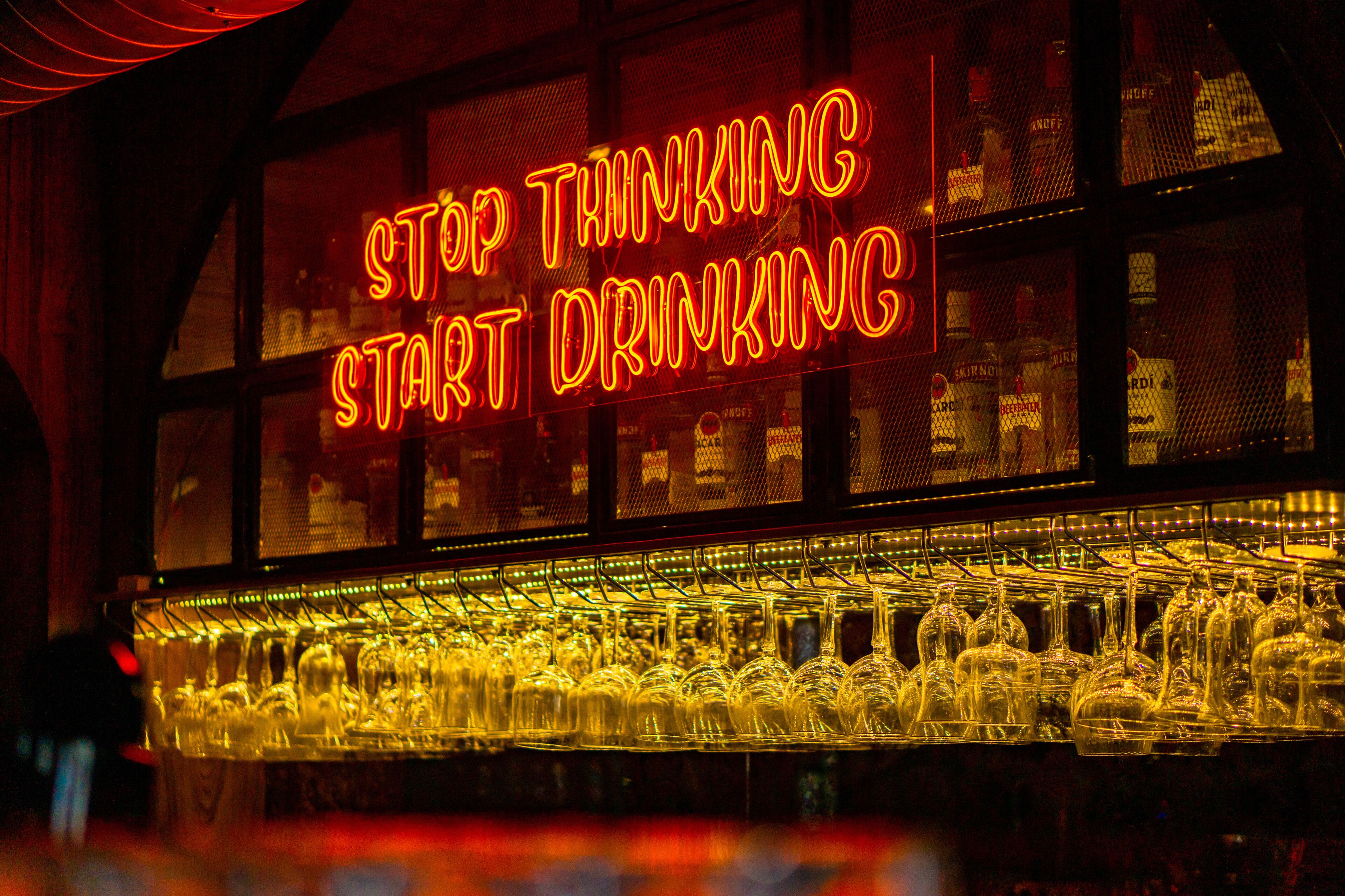 A neon sign with the text "STOP THINKING START DRINKING" above a bar area with numerous glasses.