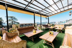 An outdoor seating area with wooden tables and benches, a glass roof, and a view of a cityscape.