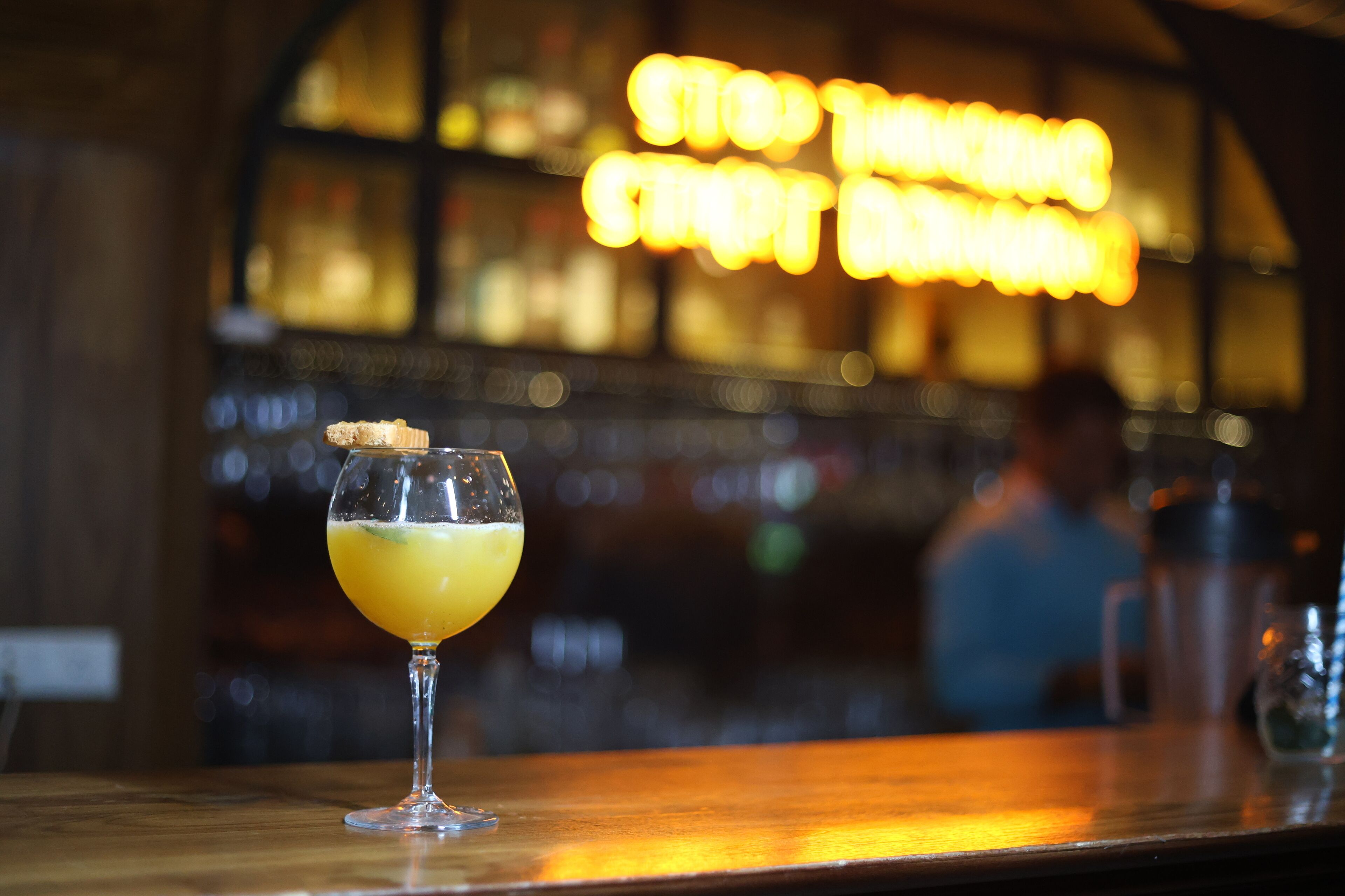 A bar setting with a glass of a yellow beverage, a cork, and a blurred background with a sign.