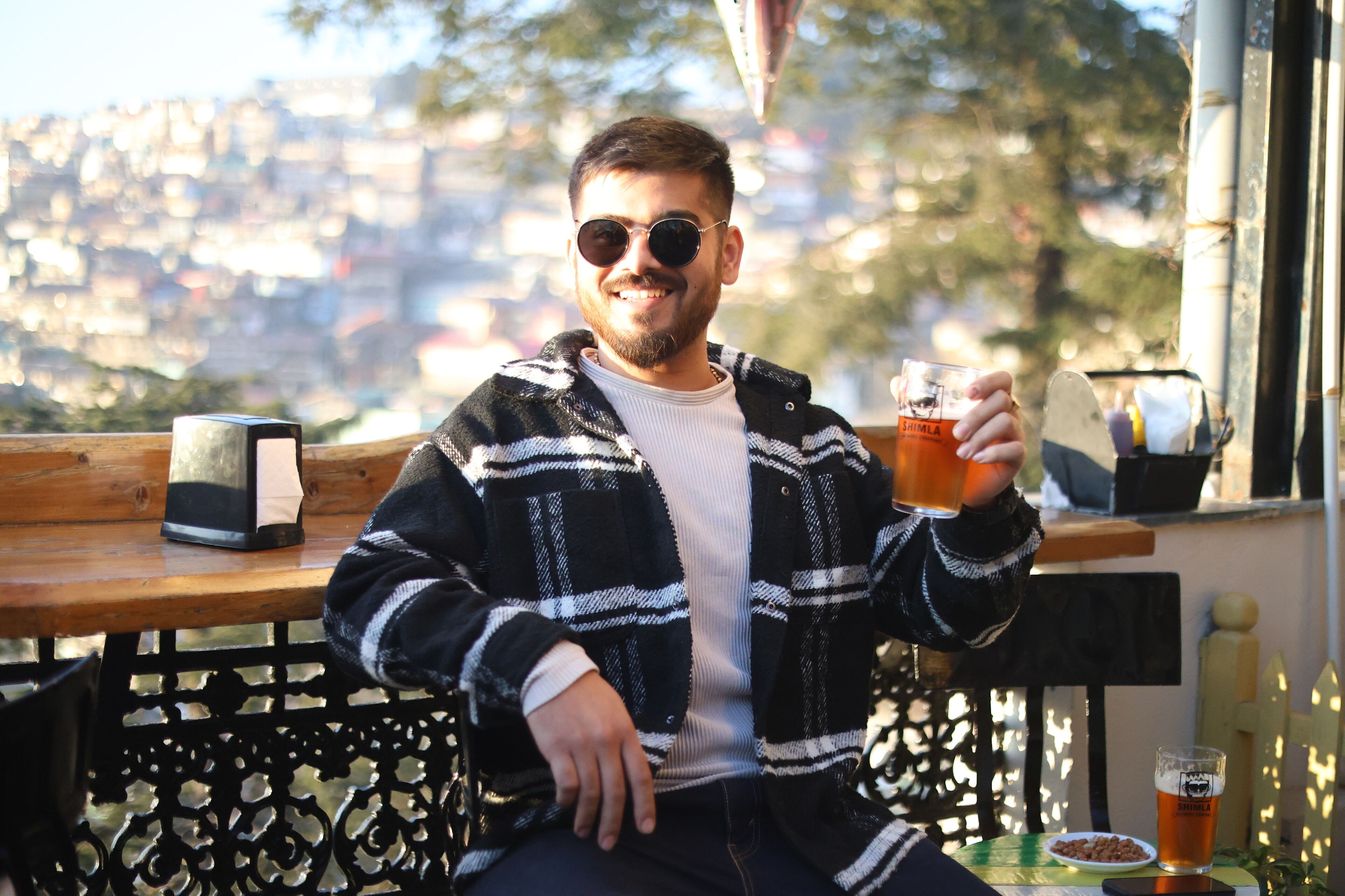 A person sitting on a bench with a drink, a bottle, and a plate of nuts on the table.
