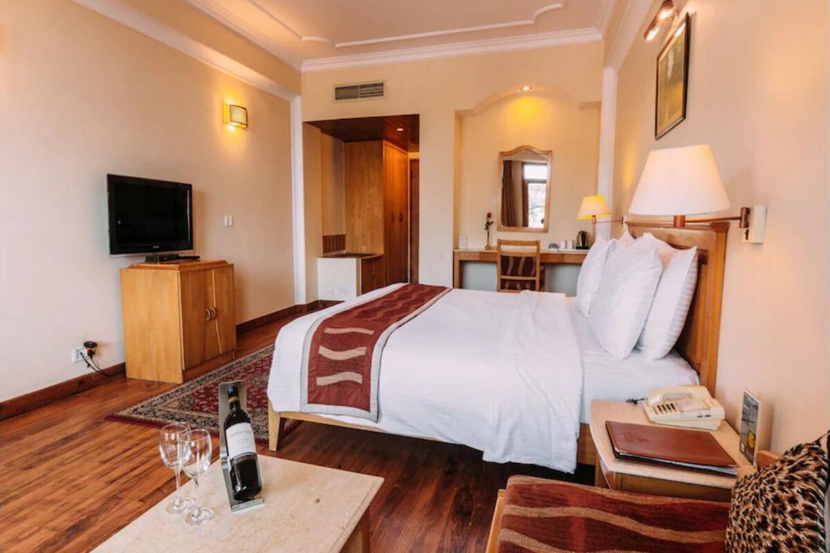 A hotel room with a bed, a television, a desk with a lamp, a chair, and a small table with a bottle and glasses.