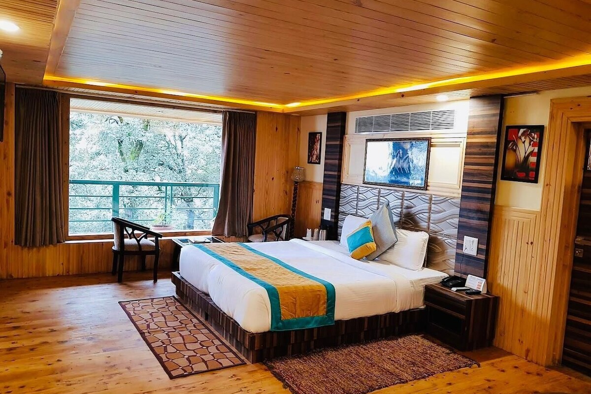 A wooden-floored bedroom with a bed, bedside table, and a view of trees through a window.