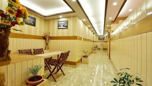 A hotel lobby with wooden paneling, a reception desk, a dining area with a table and chairs, and potted plants.