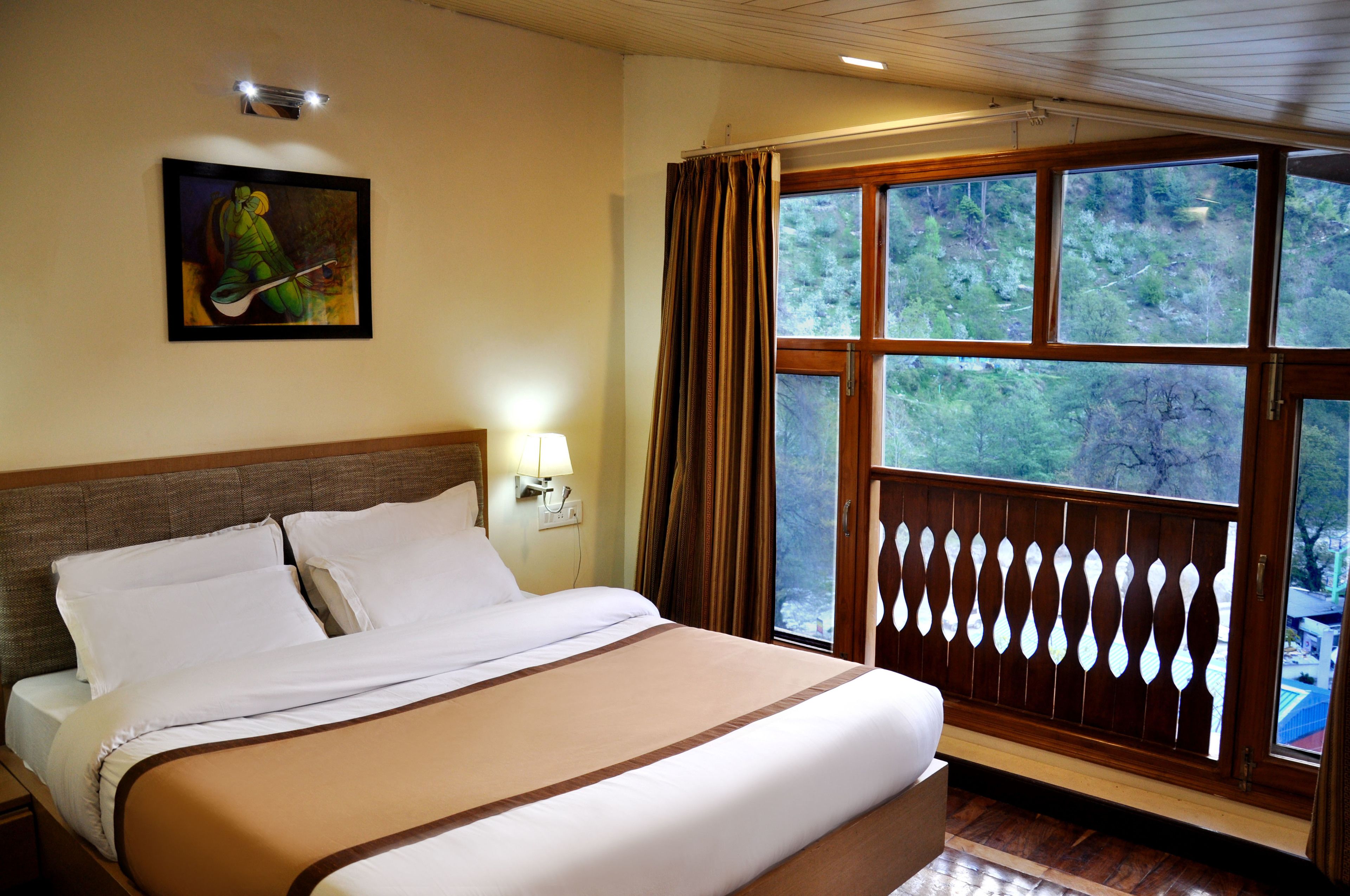 A hotel room with a large bed, a painting on the wall, a window with curtains, and a view of trees.
