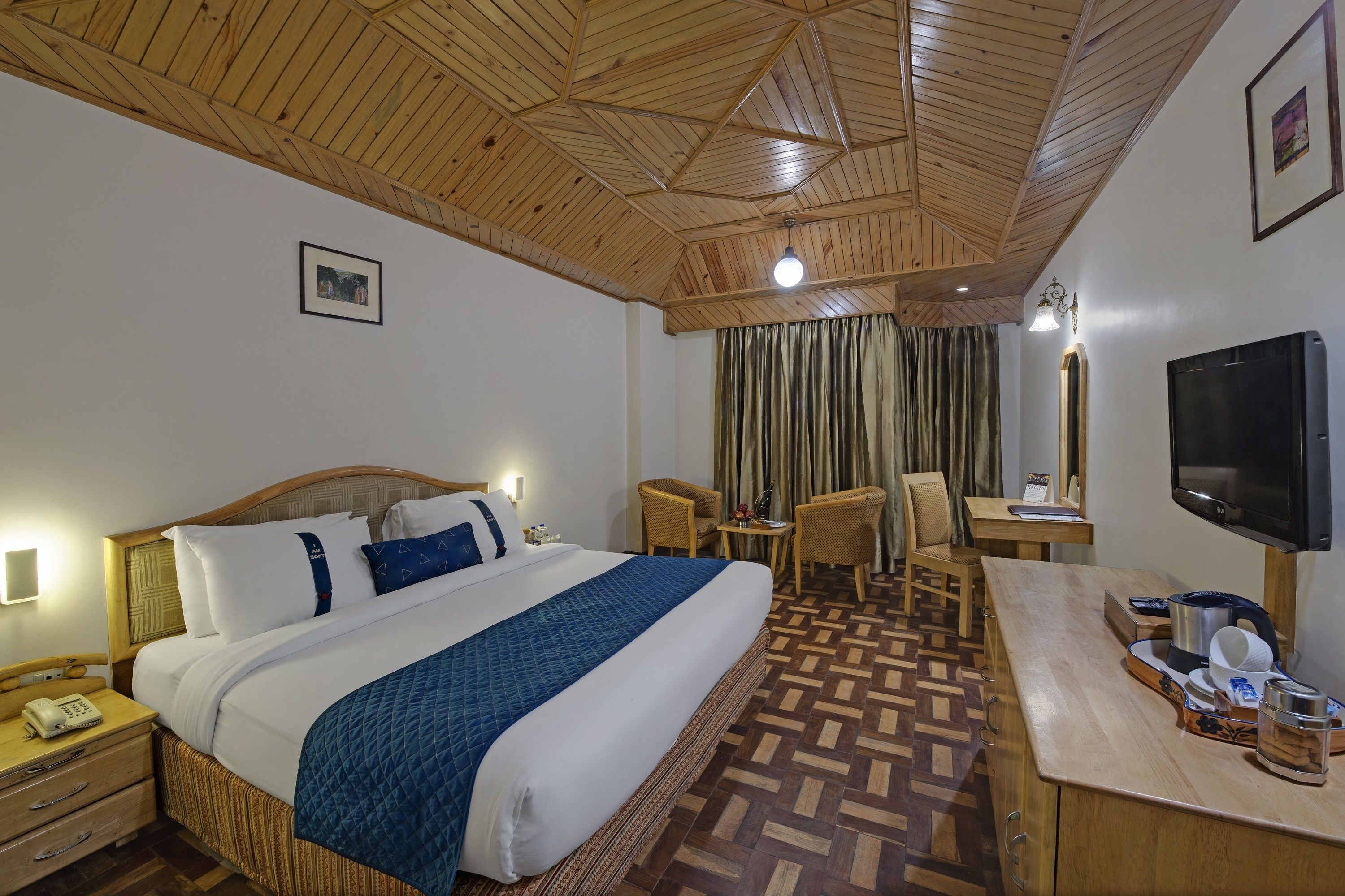 Een hotelkamer met een groot bed, houten plafond, een bureau met stoel, een televisie en een raam met gordijnen.