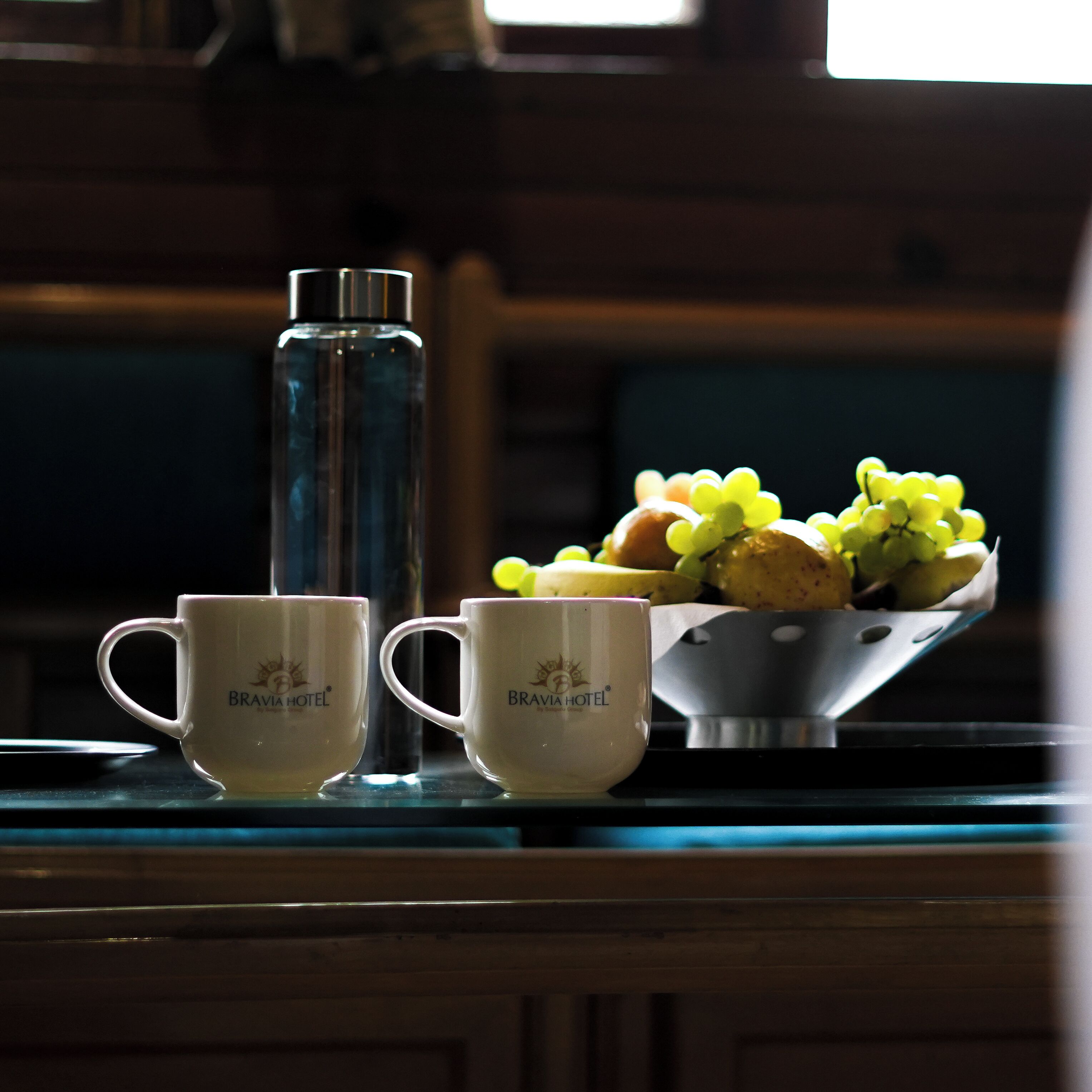 Two white cups with a logo, a clear water bottle, and a metal bowl with fruits and grapes on a wooden table.
