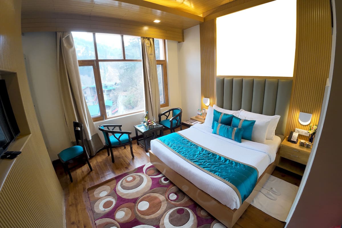A hotel room with a bed, a desk, a chair, a TV, and a window with a view of houses.