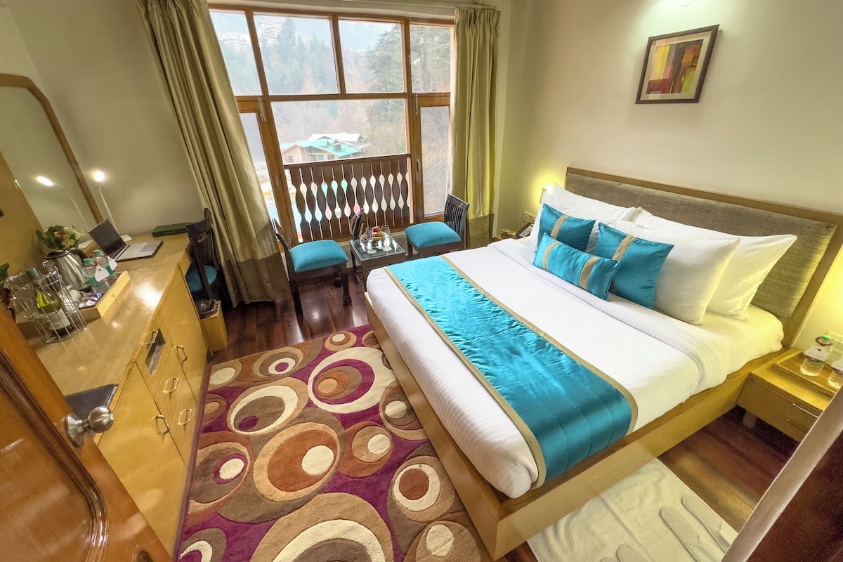 A hotel room with a large bed, a desk with a laptop, a chair, and a view of a mountainous area through the window.