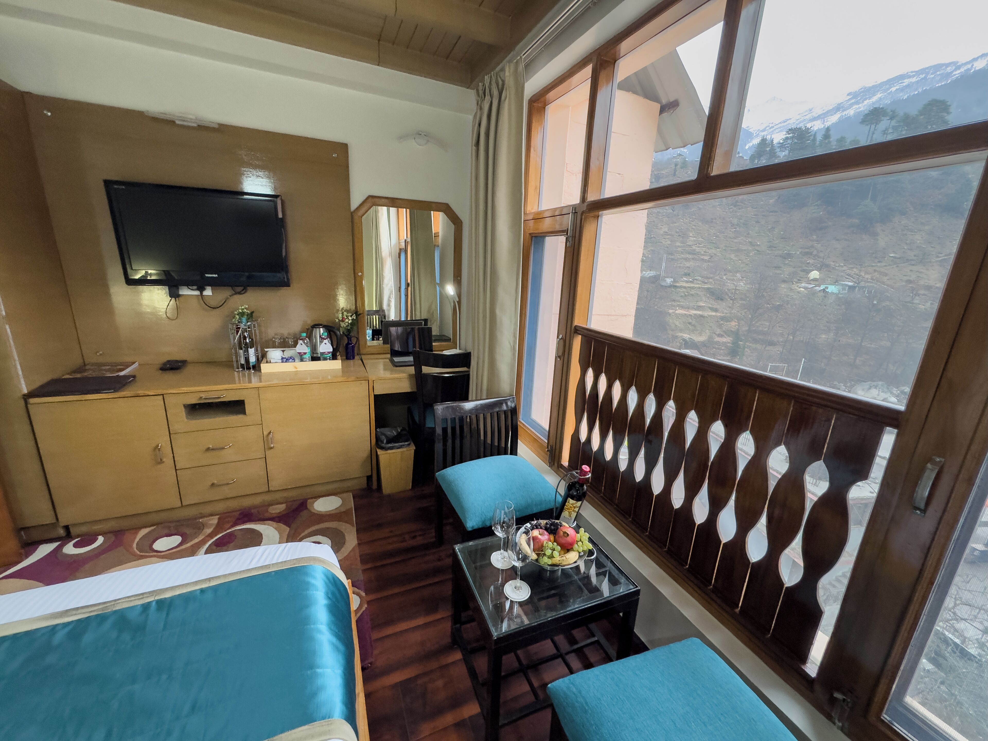 A hotel room with a bed, a desk with a computer, a TV, and a balcony with a view of mountains.