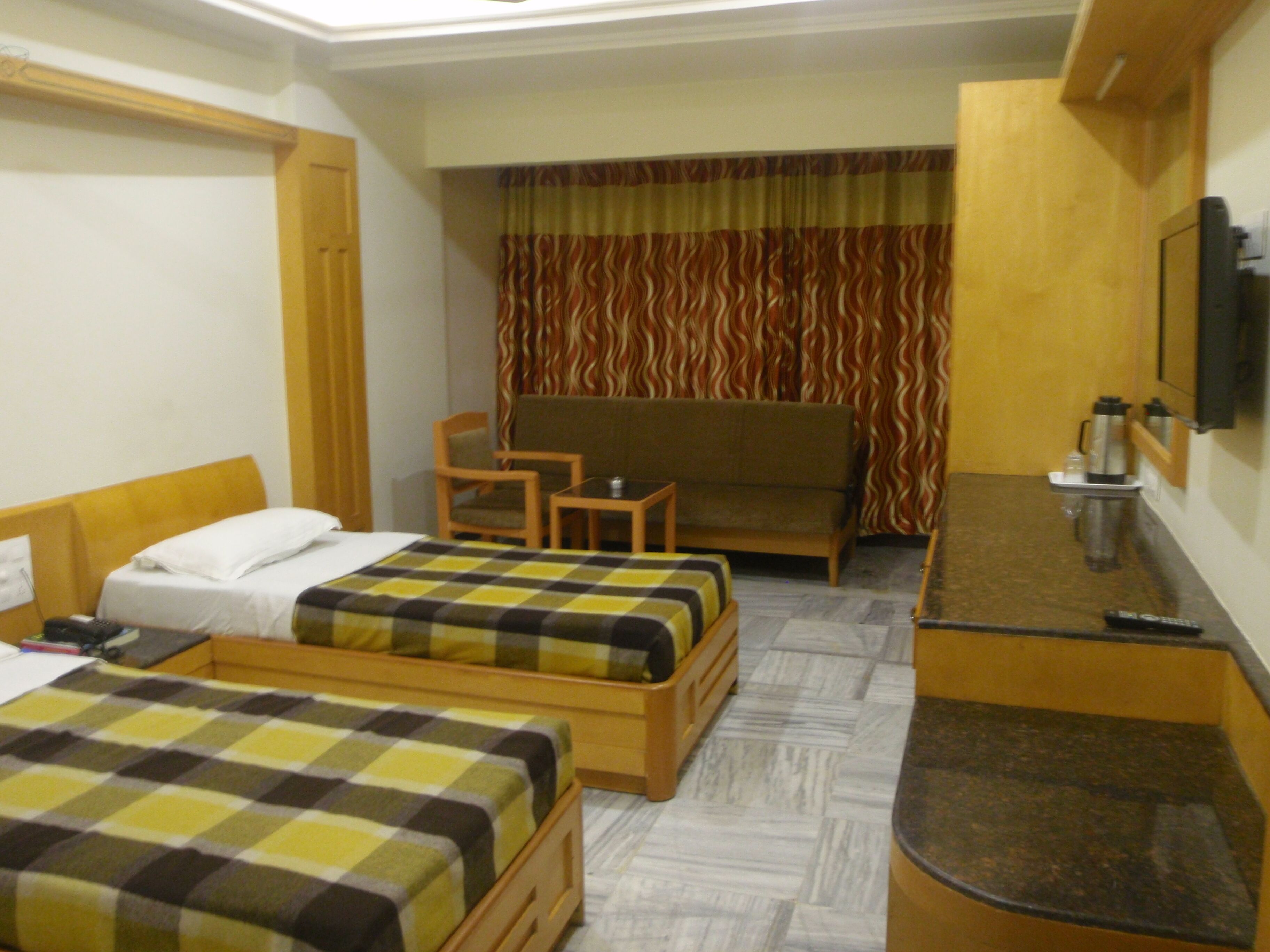 Habitación de hotel con dos camas individuales, una mesita, una silla, un sofá y un televisor en la pared.