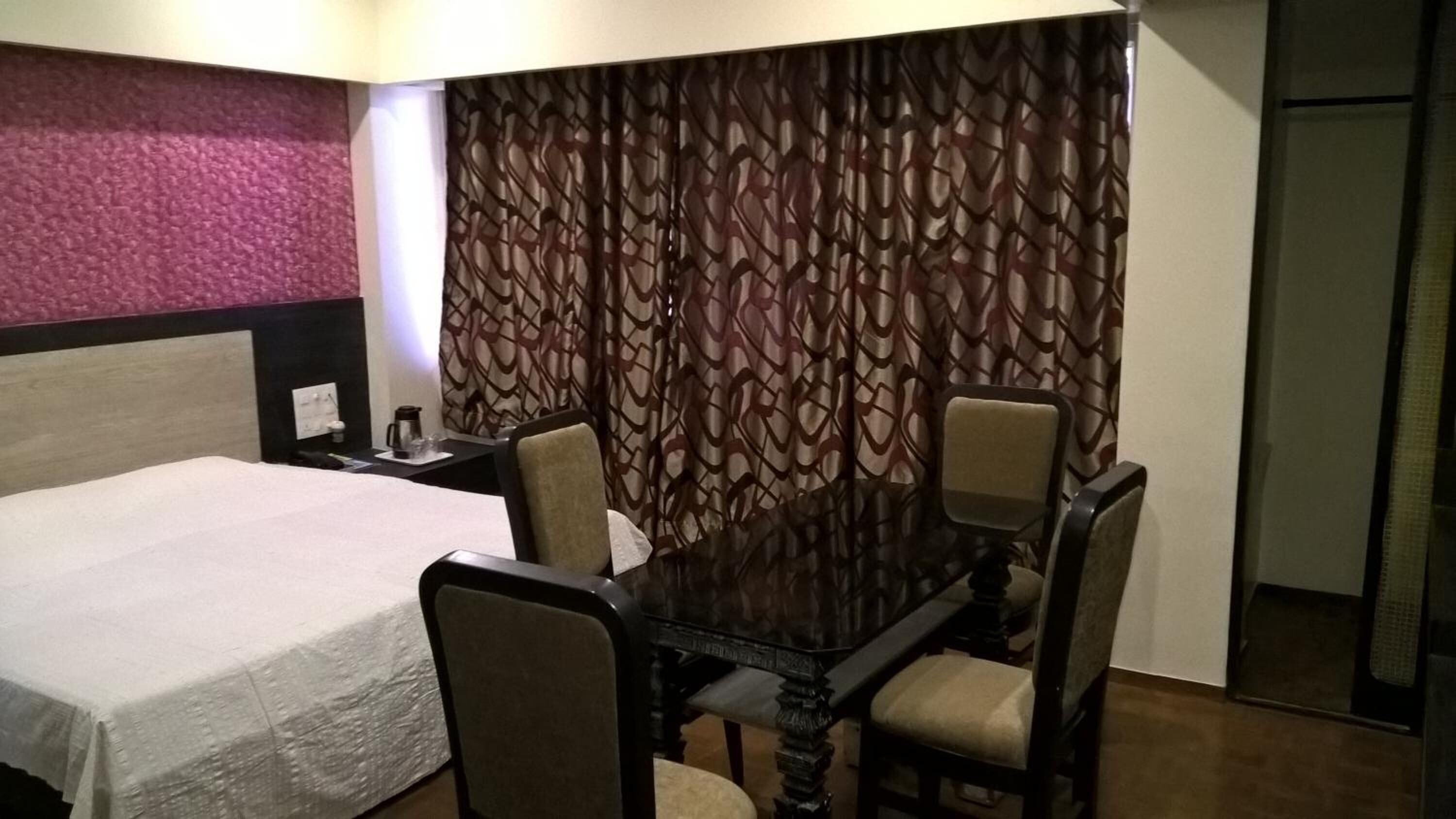 A hotel room with a bed, a dining table with four chairs, a coffee maker, and a patterned curtain.