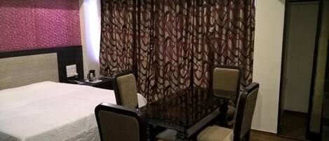 A hotel room with a bed, a dining table with four chairs, a coffee maker, and a patterned curtain.