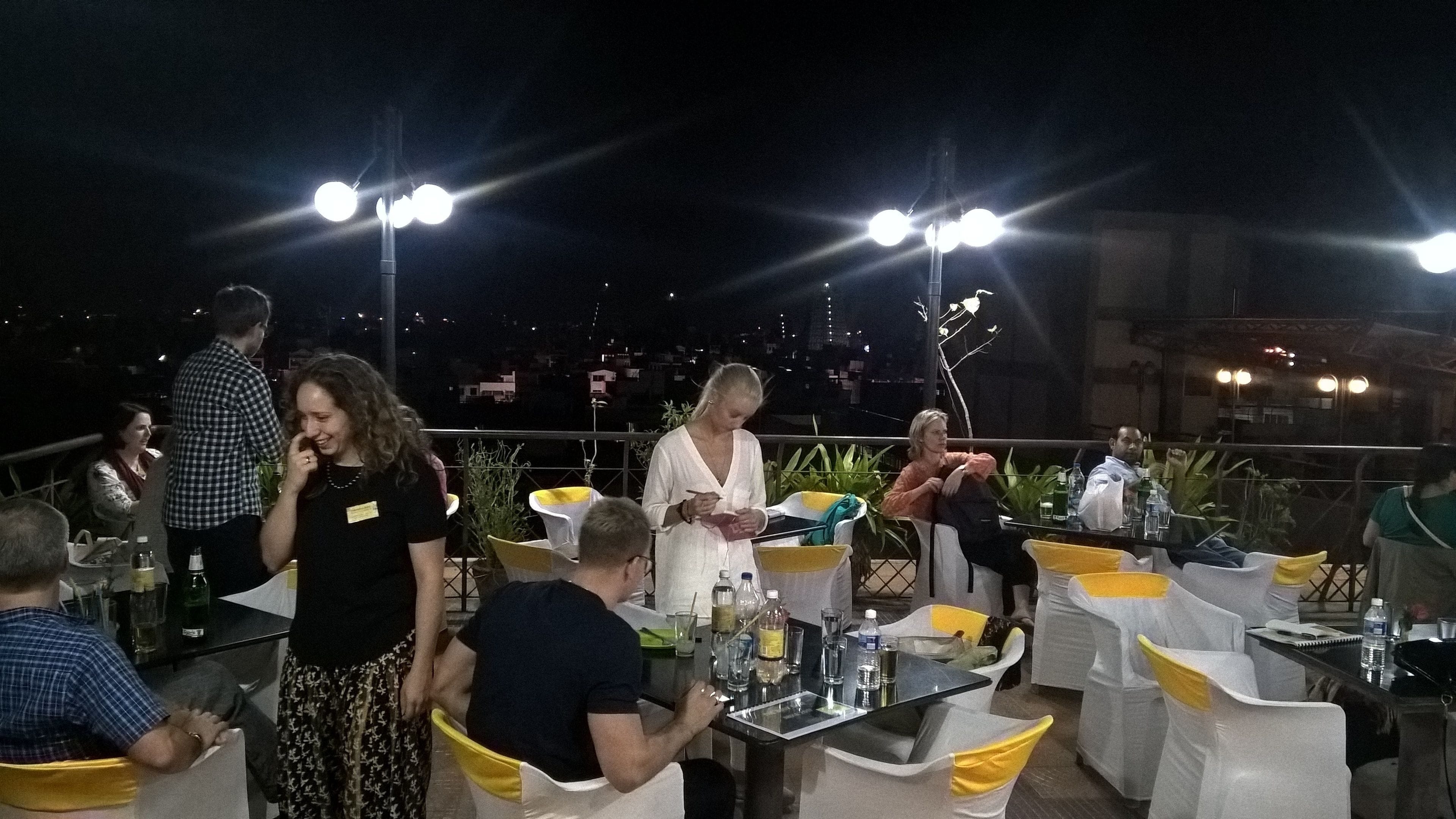 An outdoor dining area with tables and chairs, surrounded by plants and illuminated by overhead lights.