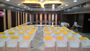 A conference room with rows of white chairs with yellow sashes, a stage with a table and chairs, and a large window with curtains. - Hotel Supreme (Madurai)