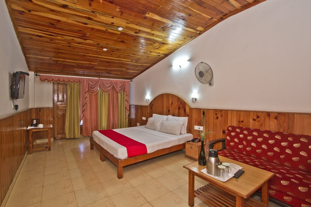 A wooden-paneled room with a bed, a sofa, a table with a kettle, and a fan on the wall.