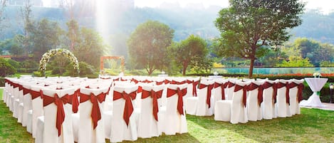 Rows of white chairs with red sashes, arranged on a grassy lawn with trees and a clear sky in the background.