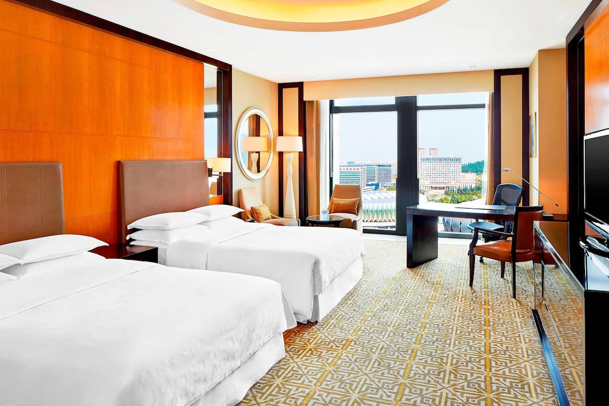 A hotel room with two beds, a desk, a chair, a TV, and a large window with a city view.