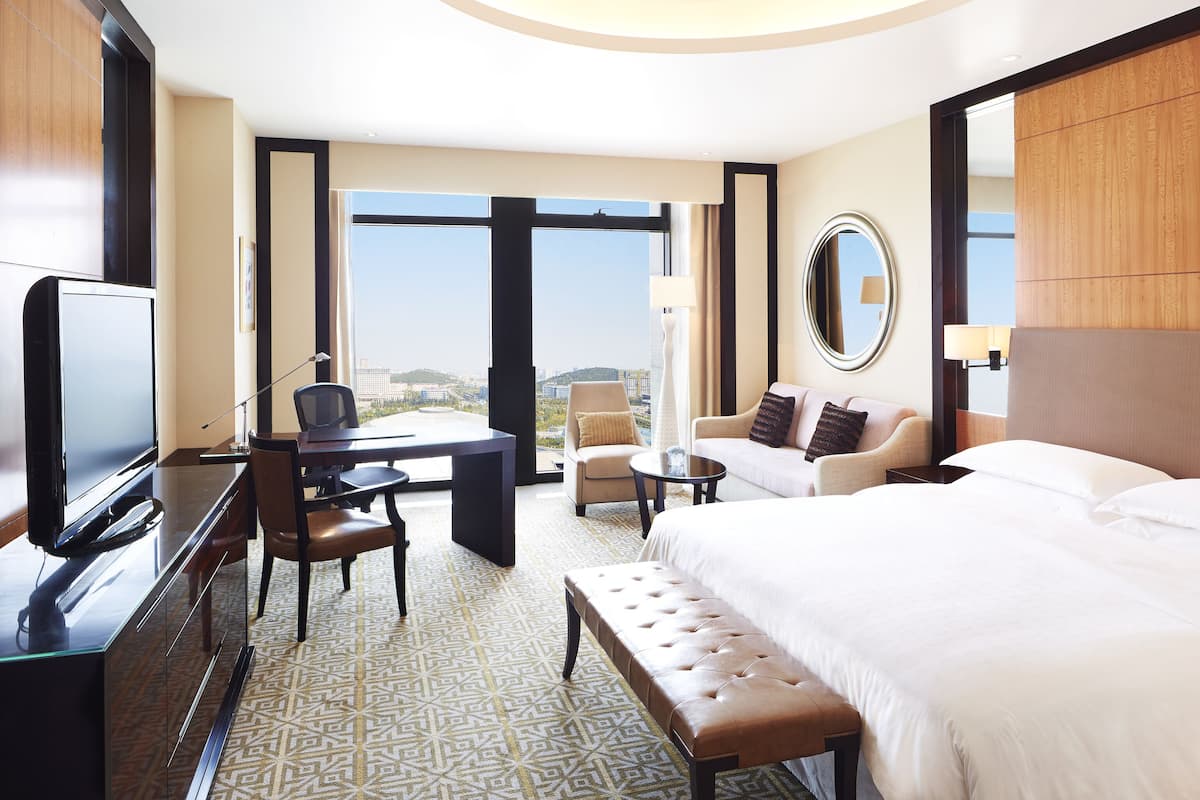 A hotel room with a large bed, a desk with a TV, a chair, a small table, and a view of the city.