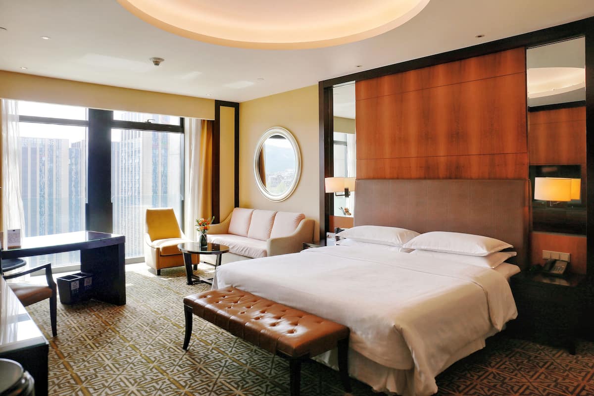 A hotel room with a large bed, a desk, a chair, a bench, a round mirror, and a view of the city through large windows.