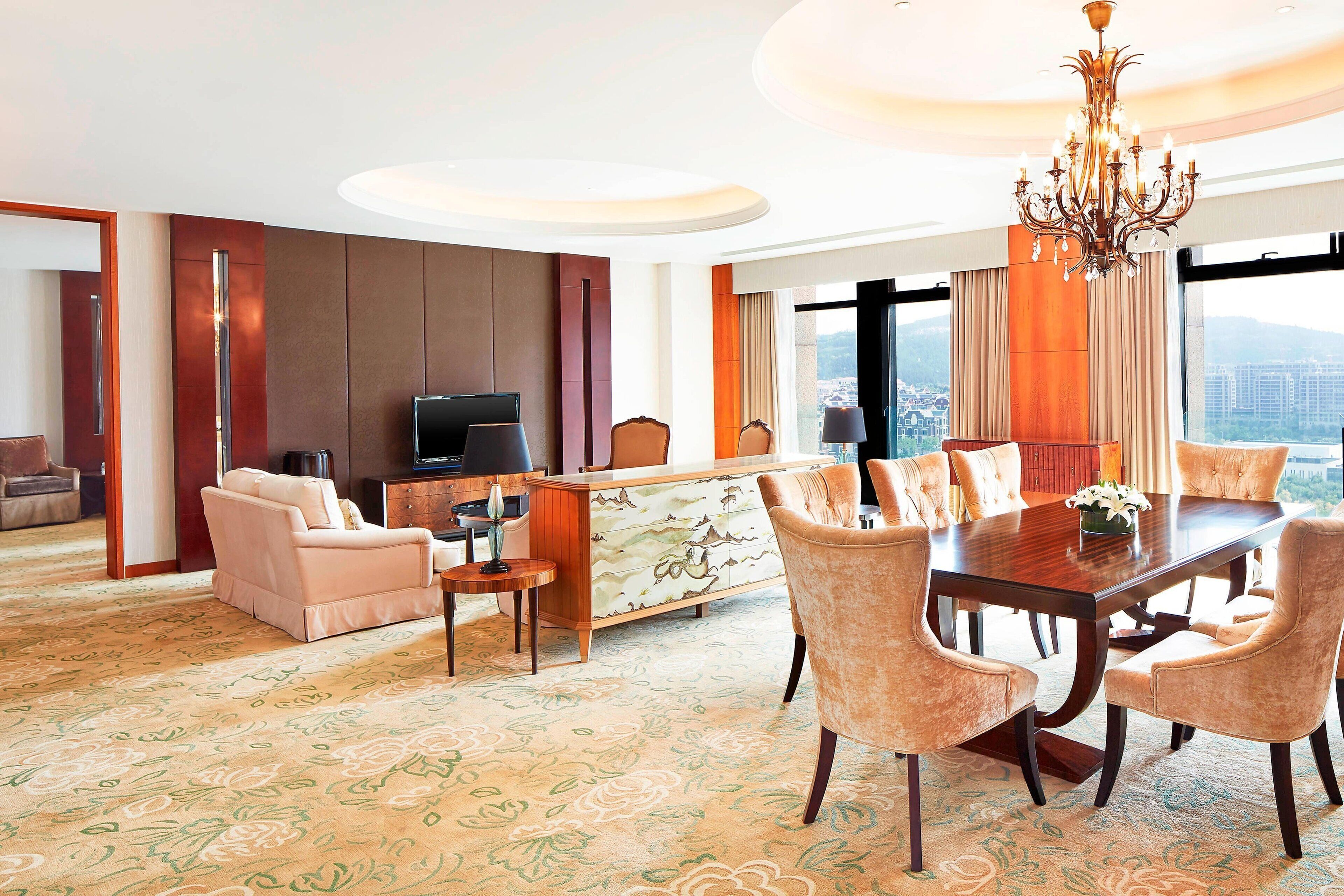 A hotel room with a dining area, a sofa, a TV, and a view of the city.