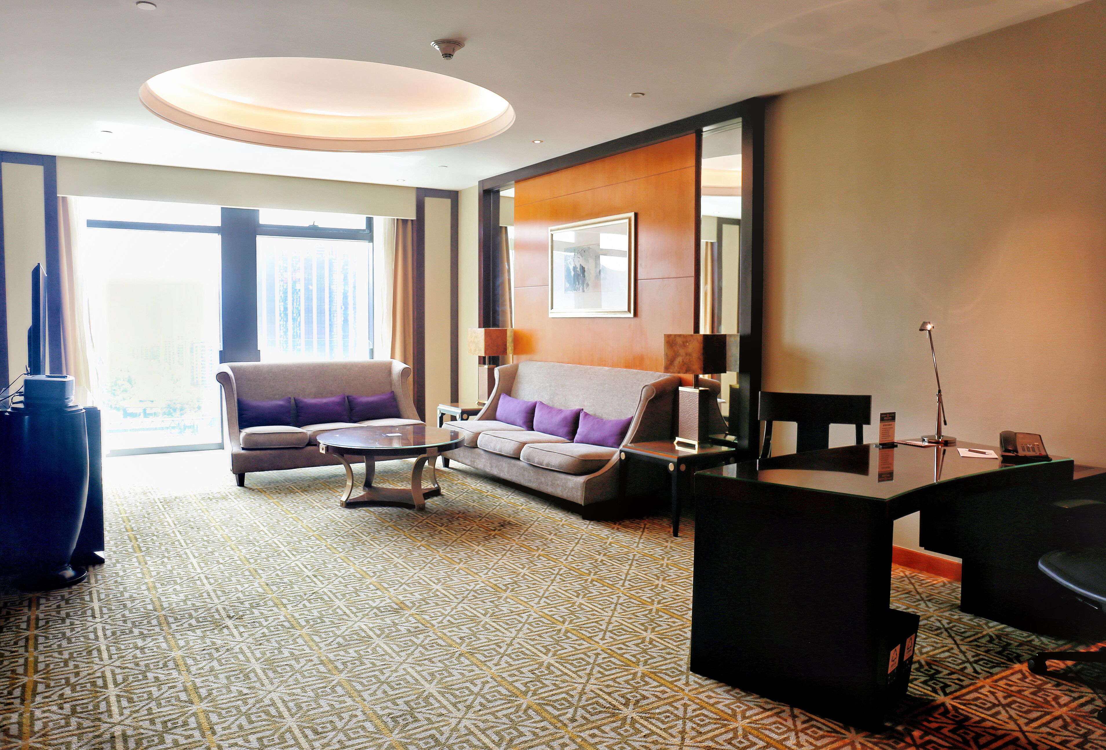A hotel room with a sofa, armchair, desk, and a large window.