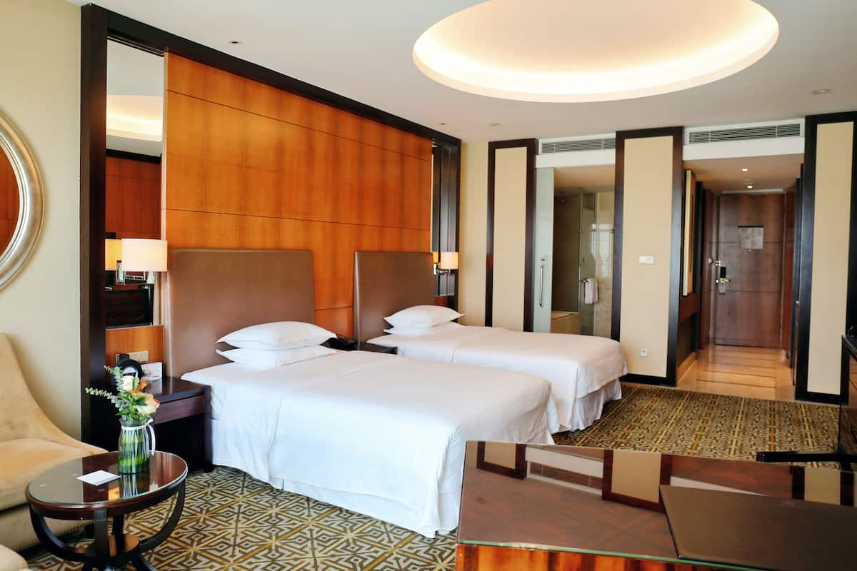 A hotel room with two beds, a sitting area with a table and a chair, a mirror, and a large mirror on the wall.