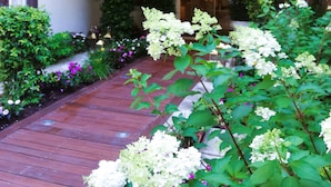 A courtyard with a wooden pathway, lush greenery, and white flowers.
